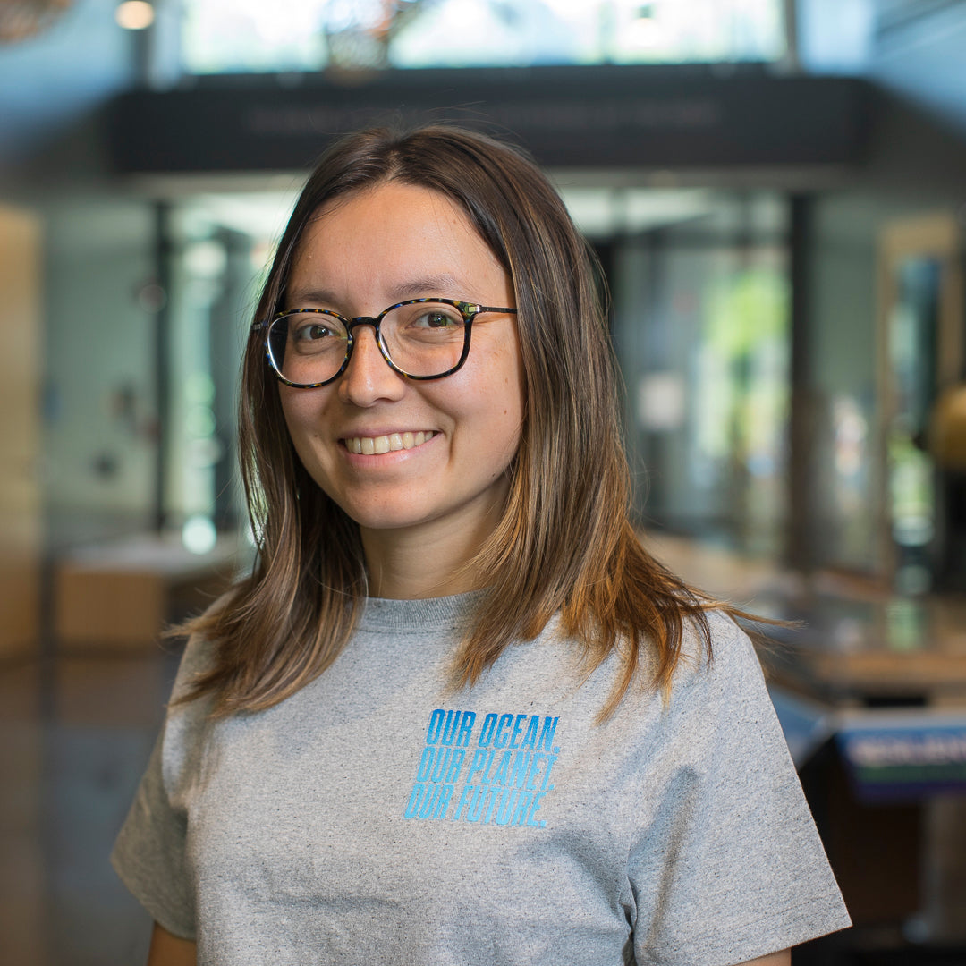 A smiling woman with glasses and shoulder-length brown hair wearing a heather gray t-shirt with a light blue graphic that reads 'OUR OCEAN OUR FUTURE' on the chest, standing in a brightly lit indoor environment.
