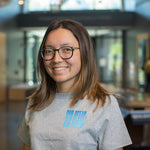 A smiling woman with glasses and shoulder-length brown hair wearing a heather gray t-shirt with a light blue graphic that reads 'OUR OCEAN OUR FUTURE' on the chest, standing in a brightly lit indoor environment.