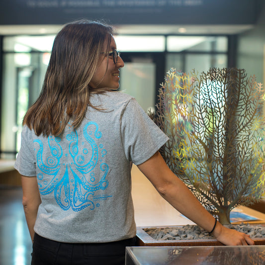 A woman with glasses and brown hair is seen from behind, wearing a light gray t-shirt with a light blue octopus graphic on the back. She is standing indoors next to a decorative metallic coral display.