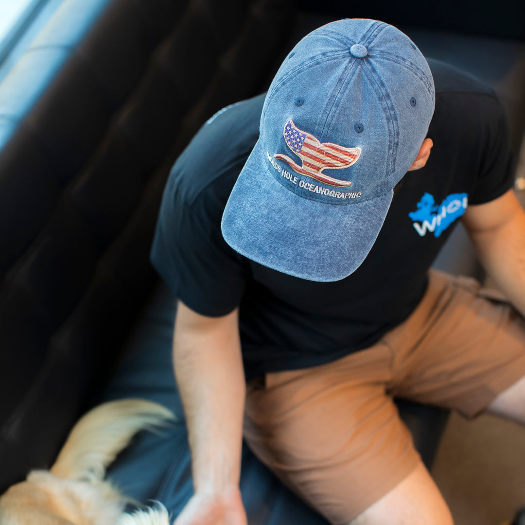 A person photographed wearing a blue denim baseball cap with an embroidered whale tail in an American flag design and the text WOOD HOLE OCEANOGRAPHIC on the front. 