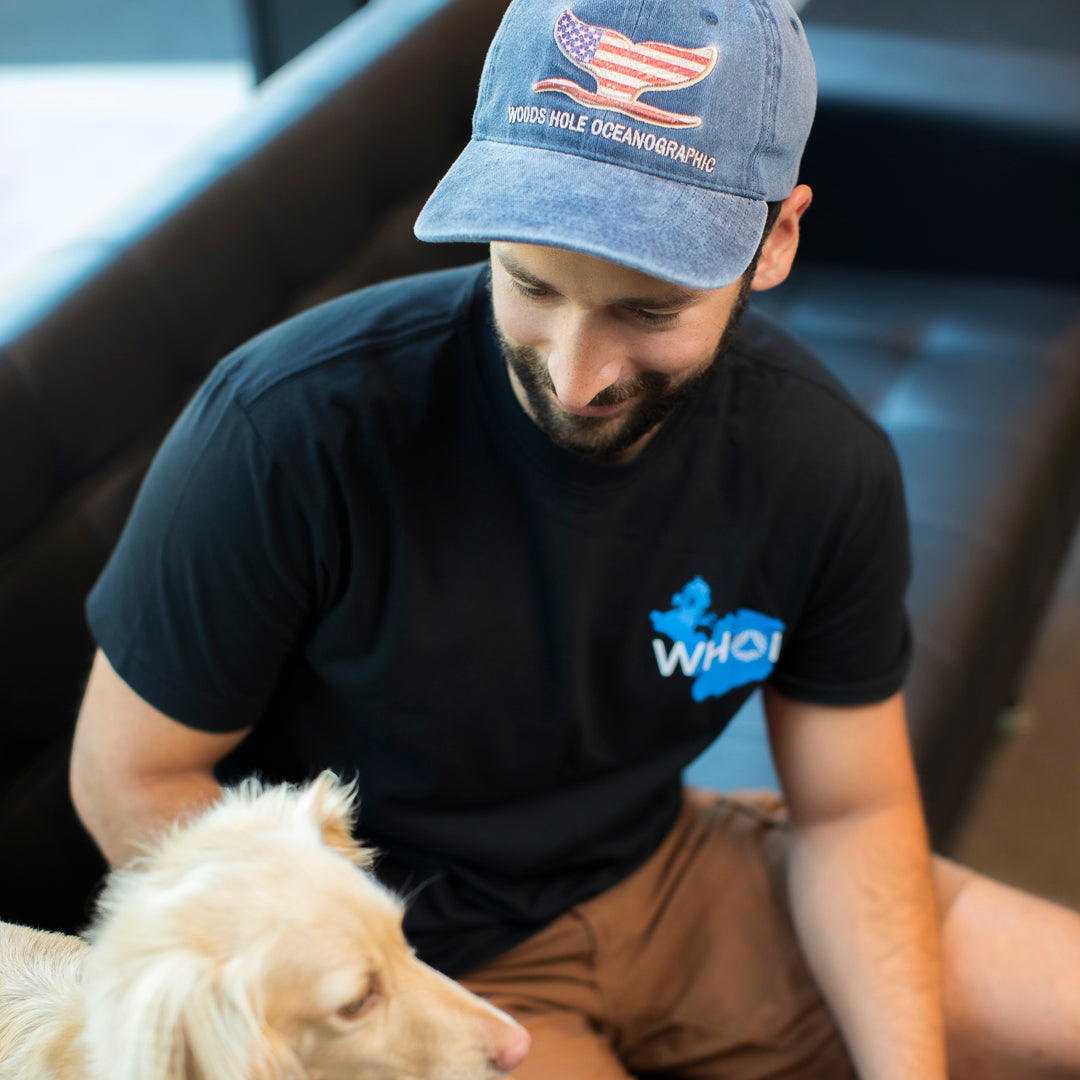 A man photographed wearing a blue denim baseball cap with an embroidered whale tail in an American flag design and the text WOOD HOLE OCEANOGRAPHIC on the front.  The man is petting a dog and sitting on a sofa.