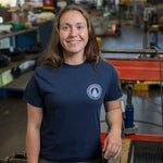 Woman wearing a navy blue t-shirt with a white WHOI Classic Seal Logo on the front left chest in an industrial work area.