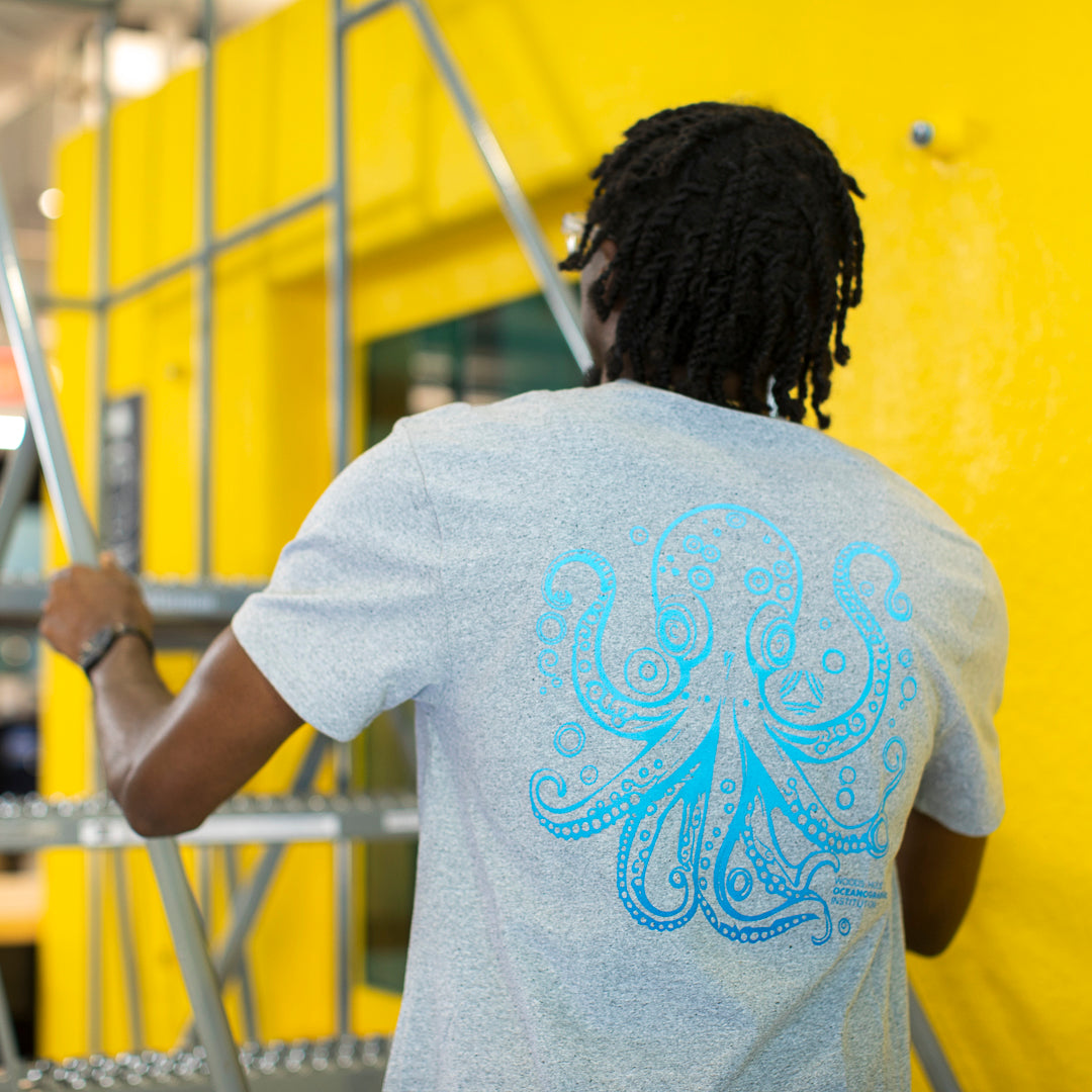 A man viewed from behind wearing a light gray heather t-shirt with a bright blue octopus graphic on the back, standing in front of a bright yellow wall.