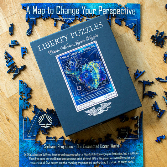 A square blue box for 'Liberty Puzzles' wooden jigsaw puzzles, featuring a colorful Spilhaus Projection map on the front cover, photographed on a wooden surface with puzzle pieces scattered around the edges. The title reads, "A Map to Change Your Perspective." 