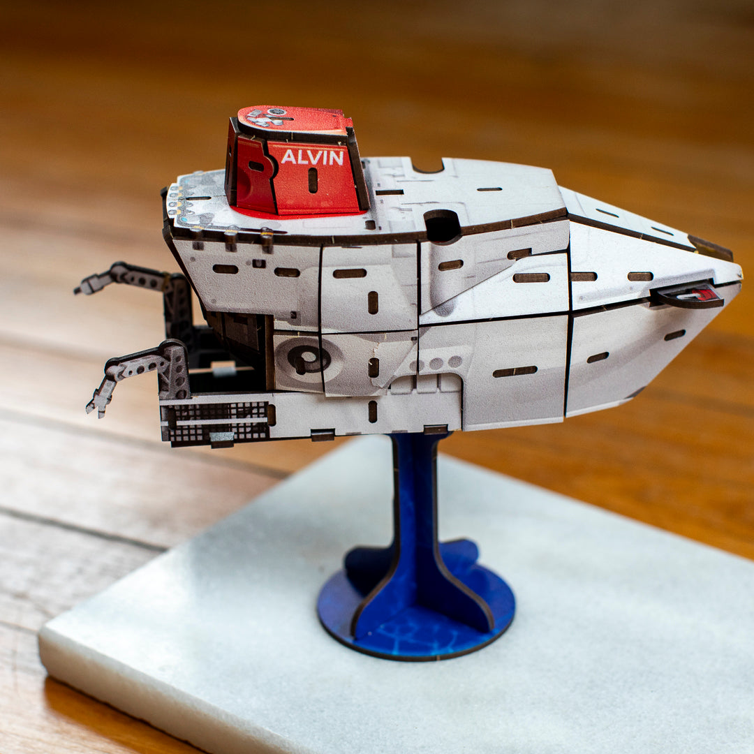 A close-up of a 3D model of the deep-sea submersible Alvin on a blue display stand. The background shows a blurred instruction booklet on a wooden table.