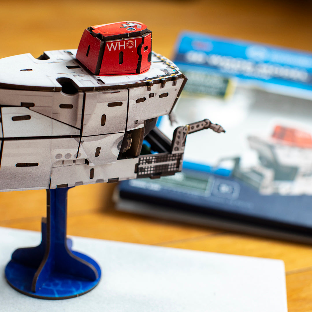 A close-up of a 3D model of the deep-sea submersible Alvin on a blue display stand. The background shows a blurred instruction booklet on a wooden table.
