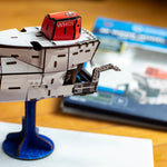 A close-up of a 3D model of the deep-sea submersible Alvin on a blue display stand. The background shows a blurred instruction booklet on a wooden table.