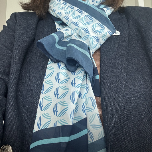 A close-up of a patterned scarf draped around a person's neck, styled with a dark blazer and brown knit sweater. The scarf features a white base with a repeating blue and teal WHOI iconepattern and solid blue borders.