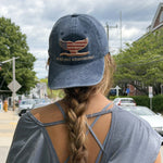 A person photographed from behind wearing a blue denim baseball cap with an embroidered whale tail in an American flag design and the text WOOD HOLE OCEANOGRAPHIC on the front. 