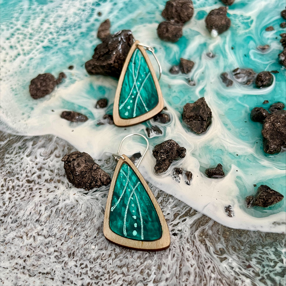 Two triangular wooden dangle earrings with a vibrant green and turquoise inlay featuring white wavy lines and dots, displayed on a textured background resembling ocean waves and dark rocks.